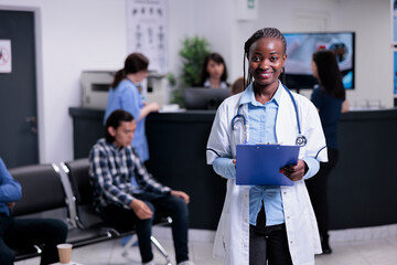 Portrait of smiling doctor with stethoscope in busy hospital reception waiting for next patient appointment. African american medic posing confident in lab coat and holding clipboard.