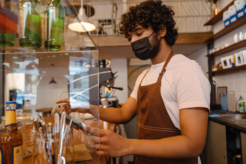 Fototapeta premium Young indian curly barista in face mask working with tablet