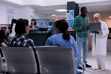 Older woman medic holding laptop with mri scan results talking with african american patient in private clinic waiting room. Senior doctor having conversation with younger man checking appointment.