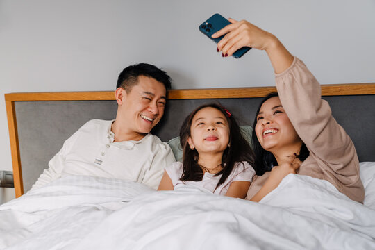 Asian Family With Little Girl Taking Selfie Photo While Lying In Bed