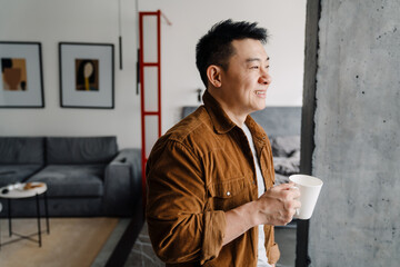 Brunette adult asian man smiling and drinking coffee at home
