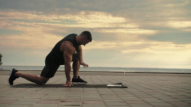 Bodybuilder shows a stretching exercise, training online, through a computer against a background of the sea. Training on the street.