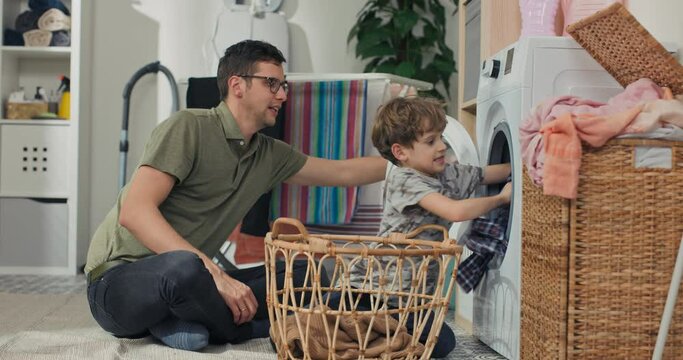 Father spends time with young children in laundry room, bathroom, performs household duties while on paternity leave, takes care of son, shows boy how to operate washing machine turn on program.