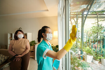 Asian young woman in protective mask and gloves wiping window with rag with owner controlling the work in background