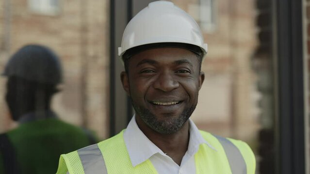 Portrait of african american foreman posing at residential construction site. Successful builder contractor in white hardhat looking at camera with sincere smile on face.