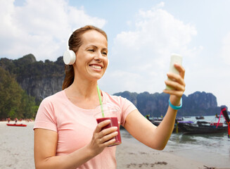 travel, tourism and vacation concept - woman in earphones with takeaway smoothie drink in plastic cup and smartphone listening to music over tropical beach background in french polynesia