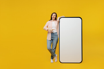 Full body young woman she 30s in striped shirt white t-shirt point index finger on big huge blank screen mobile cell phone with workspace copy space mockup area isolated on plain yellow background