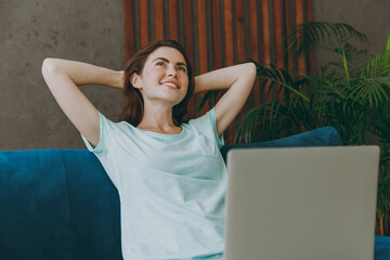 Young minded fun woman she 20s in casual clothes mint t-shirt hold hands behind neck look aside use work on laptop pc computer study online sit on blue sofa indoor rest at home in own room apartment