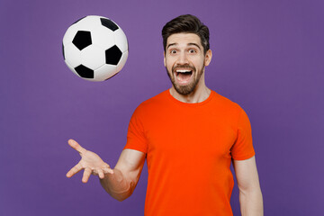 Young cheerful cool surprised excited satisfied fun fan man he wears orange t-shirt cheer up support football sport team watch tv live stream toss up soccer ball isolated on plain purple background.