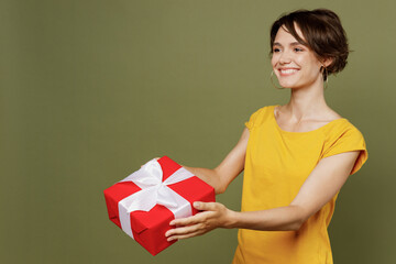 Young smiling happy cheerful woman she 20s wear yellow t-shirt hold in hand give red present box with gift ribbon bow isolated on plain olive green khaki background studio People lifestyle concept.