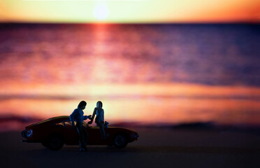 Miniature lover couple stand beside of classic car with a background of sunset sea beach