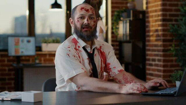 Ugly dirty office zombie sitting at desk while working on modern laptop. Frightening creepy monster with deep and bloody wounds and scars using portable computer for work in company workspace.