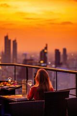 Young woman with glass of wine rest at luxury rooftop restaurant watching orange sky sunset. Female...