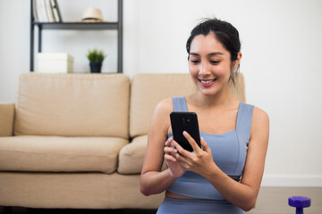 Lifestyle Attractive asian young fitness woman holding smartphone relaxing after workout at home. Smiling female using cell phone checking newsfeed on social media while exercise
