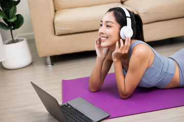 Young beautiful asian woman put on headphone listening music by laptop relax after workout. Lay down on yoga mat. Cheerfully sporty female workout and exercise wearing sport wear at home.