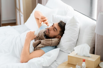 Obraz na płótnie Canvas people and health problem concept - unhappy sick man spraying his nose with nasal spray lying in bed at home