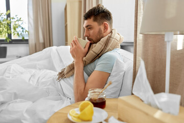 Fototapeta na wymiar health, illness and people concept - sick man drinking hot tea with lemon and honey in bed at home