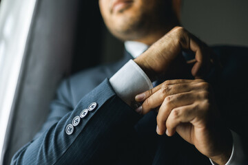 Confident businessman buttoning or adjust classic blue suit near window in hotel room at the morning. Handsome man wearing a nice suit on wedding day.