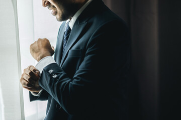 Confident businessman buttoning or adjust classic blue suit near window in hotel room at the morning. Handsome man wearing a nice suit on wedding day.