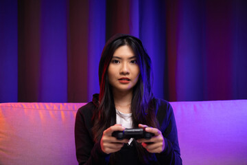 Playing video game. Young asian pretty woman sitting on sofa holding joystick in living room. Happy female Professional Streamer chinese wearing hoodie playing game online in dark room neon light.