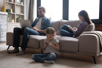 Young family of three holding and using different electronic devices while sitting on sofa in the living room