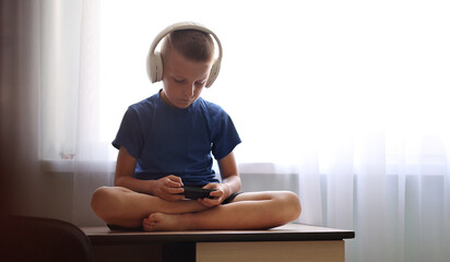 Fototapeta na wymiar Child's dependence on a mobile device theme. Child with headphones on neck is sitting on table and uncontrollably playing online games on smartphone. children's addiction to online games. virtual life