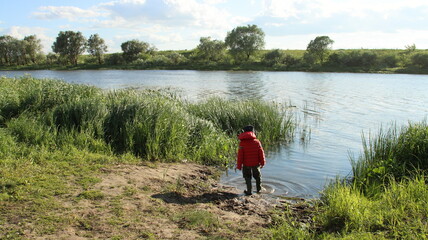 A boy in a red jacket and a blue hat stands on the swampy bank of the river