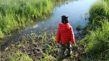 A boy in a red jacket and a blue hat stands on the swampy bank of the river