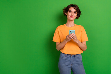 Photo of adorable dreamy young lady wear orange outfit looking empty space chatting modern gadget isolated green color background