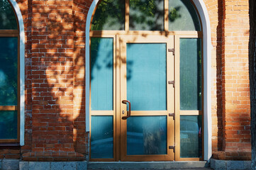 Entrance door made of glass and plastic in an old red brick building. Reflections and shadows of tree foliage on a summer morning. Blinds behind glass. Arched modern designs. Reinvention concept.
