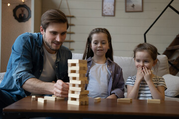 Joyful father with daughters plays at home. Kids remove wooden blocks from tower. Board game.