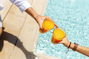 Top view hands of couple in casual wear sitting poolside together and drinking cocktails or orange juice. Carefree relax at the pool. Vocation concept.