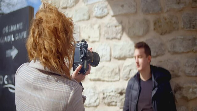 A woman takes a picture of a guy in front of a wall. ACTION. Red-haired woman photographer looks at the man. The process of photographing a man
