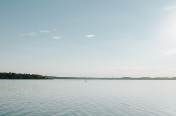 Lake, forest on horizon, unrecognizable person riding SUP board on water. Summer natural landscape, wide view