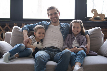 Little daughters sitting next to daddy at home smile look at camera. Celebrating Happy Father Day concept