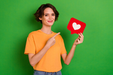 Photo of charming pretty young lady wear orange outfit pointing heart like sign isolated green...