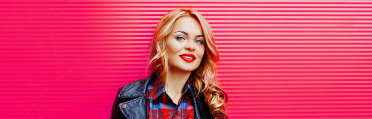 Portrait of beautiful blonde young woman with red lipstick wearing black rock jacket on pink background