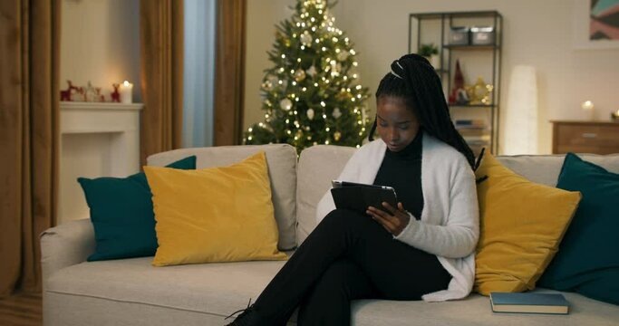 An African-looking student sits on sofa, holding tablet and ordering Christmas presents for friends. The girl is dressed in black clothes and white cardigan. There are pillows and book on the sofa.
