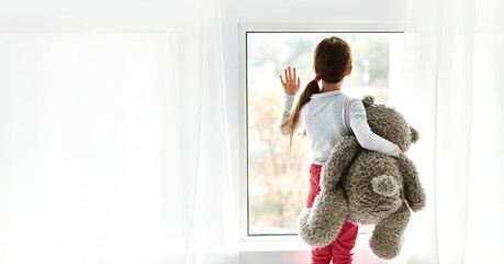 Loneliness. Lonely orphan girl in an orphanage looking outside while holding teddy bear the window. Child feelings concept - obrazy, fototapety, plakaty