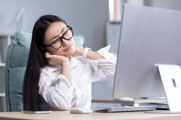 Overtired Asian woman office worker, has severe neck pain, massages neck muscles with hand, female worker in office at computer.