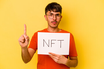 Young hispanic man holding nft placard isolated on yellow background showing number one with finger.