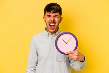 Young hispanic man holding a clock isolated on yellow background screaming very angry and aggressive.