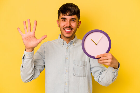 Young hispanic man holding a clock isolated on yellow background smiling cheerful showing number five with fingers.