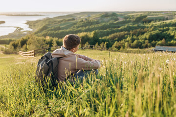 Naklejka na ściany i meble Tourist with backpack sitting on top of hill in grass field and enjoying beautiful landscape view. Rear view of teenage boy hiker resting in nature. Active lifestyle. Concept of local travel