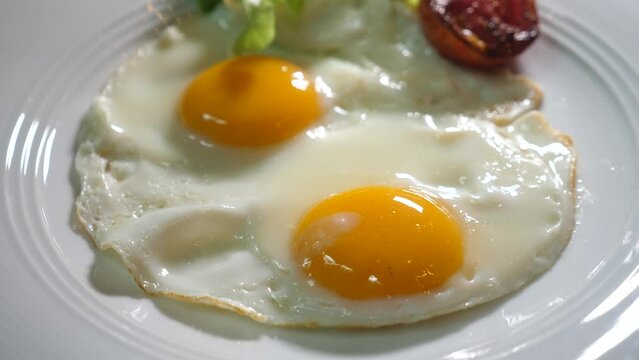 Woman hands with fork and knife cut sunny side up egg on white plate. Liquid egg yolk close-up. Concept of simple fresh healthy food on breakfast at home, fried eggs with tomato and salad leaf.