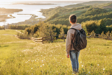 Tourist with backpack standing on top of hill in grass field and enjoying beautiful landscape view. Rear view of teenage boy hiker resting in nature. Active lifestyle. Concept of local travel