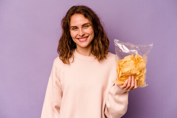 Young caucasian woman holding a bag of chips isolated on purple background happy, smiling and...