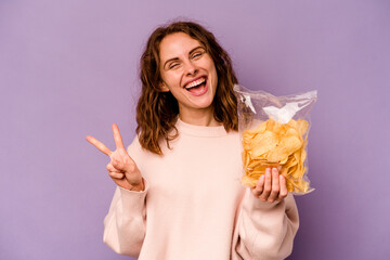 Young caucasian woman holding a bag of chips isolated on purple background joyful and carefree showing a peace symbol with fingers.