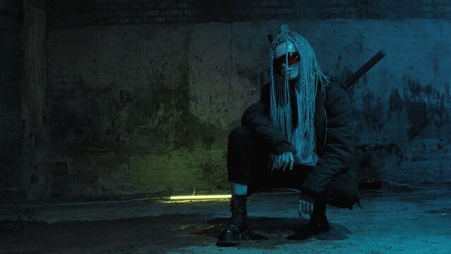 girl with dreadlocks and katanas in red glasses posing against a neon brick wall