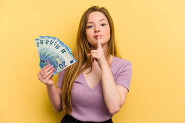 Young caucasian woman holding banknotes isolated on yellow background keeping a secret or asking...